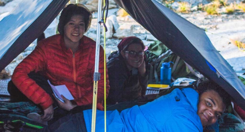Three people resting under a tarp shelter smile for the photo. 